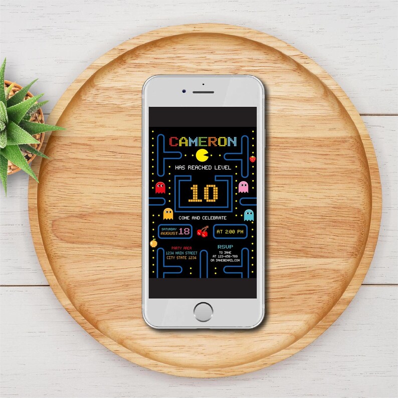 May include: A smartphone shows a birthday invitation with a Pac-Man design. The invitation includes the name "CAMERON", the number "10", and the words "COME AND CELEBRATE". The date is August 18th at 2:00 PM. The phone is on a wooden tray.