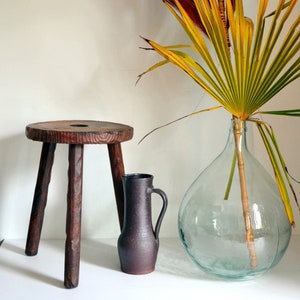 Peut inclure: Un tabouret en bois rustique à trois pieds et un dessus rond, un pichet en céramique brun avec une anse et un grand vase en verre transparent avec une fronde de palmier séchée.