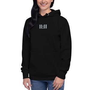 Op de afbeelding: Zwarte hoodie met een voorzak en een capuchon met trekkoord. De hoodie heeft de tekst "11:11" in lichtgrijze kleur geborduurd op de borst. Het model draagt een blauwe spijkerbroek.