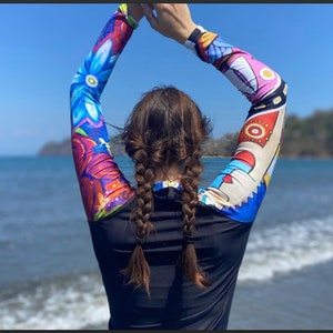 May include: A woman wearing a black long-sleeved shirt with colorful abstract designs on the sleeves. She has her arms raised above her head and is looking out at the ocean.