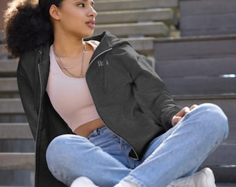 Può includere: Una donna indossa una felpa con cappuccio grigia scura con zip e la parola "HE" sul davanti. È seduta su una scalinata con jeans blu e scarpe da ginnastica bianche.