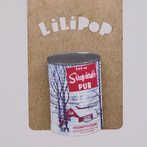 Op de afbeelding: Een rood en wit blik met een winterscène en de tekst "540 ml Sirop d'érable PUR PRODUIT DU QUEBEC REFRIGEREZ UNE FOIS OUVERT FAIT AU CANADA".