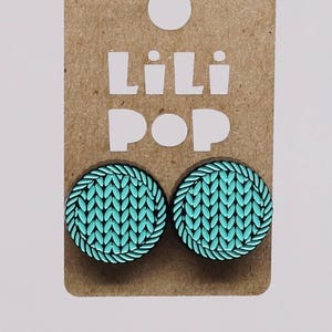 Peut inclure: Une paire de boucles d'oreilles rondes en tricot bleu sarcelle et noir. Les boucles d'oreilles sont rondes et ont une bordure noire. Les boucles d'oreilles sont sur une carte brune avec le texte "Lili Pop" imprimé dessus.