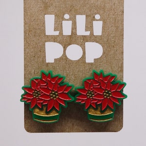 Peut inclure: Une paire de boucles d'oreilles de Noël en poinsettia rouge et verte. Les boucles d'oreilles sont en plastique et ont une bordure dorée. Les boucles d'oreilles sont sur une carte brune avec le texte "Lili Pop" imprimé dessus.