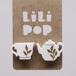 Op de afbeelding: Een paar witte theekop- en theepot-oorbellen met gouden bladontwerpen. De oorbellen worden getoond op een bruine kaart met de tekst "Lili Pop" in wit.