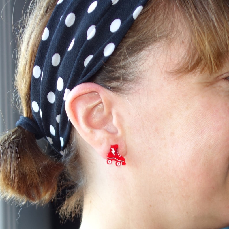 May include: Red roller skate earrings with a white lightning bolt design. The earrings are worn by a person with a black and white polka dot headband.