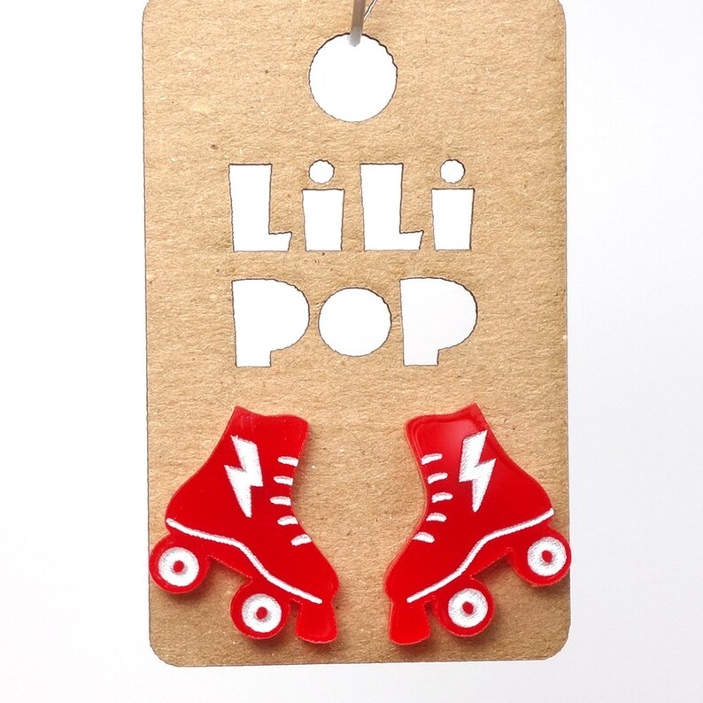 May include: Red roller skate earrings with white lightning bolt designs. The earrings are displayed on a brown card with the text "Lili Pop" in white.