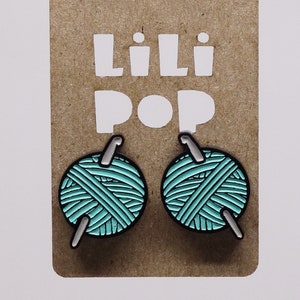 May include: A pair of teal blue yarn ball earrings with a silver knitting needle. The earrings are on a brown card with the text "Lili Pop" printed on it.