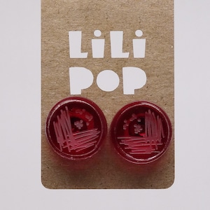 Peut inclure: Une paire de boucles d'oreilles bouton rouge avec un motif blanc. Les boucles d'oreilles sont emballées sur une carte brune avec le texte "Lili Pop" imprimé en blanc.