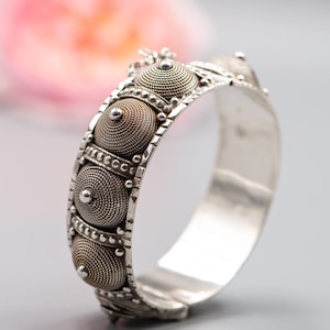 May include: A silver bracelet with detailed craftsmanship. The bracelet showcases a sequence of circular, textured components with a spiral pattern, complemented by smaller, beaded embellishments. The design is symmetrical, and the bracelet has a hinged clasp.