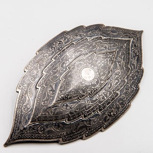 May include: A silver-coloured, leaf-shaped dish with detailed, engraved floral patterns. The dish features a layered design, with a central circular detail. The overall aesthetic is ornate and detailed, suggesting a decorative or serving piece.