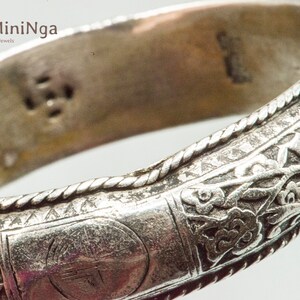 May include: Silver bangle bracelet with intricate floral designs and a circular stamp with an inscription. The bracelet has a rope-like border.