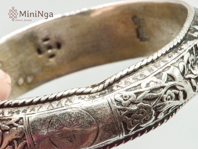 May include: Silver bangle bracelet with intricate floral designs and a circular stamp with an inscription. The bracelet has a rope-like border.