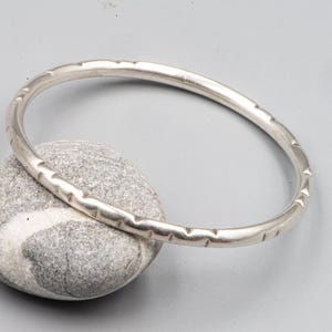 Pode incluir: Uma pulseira rígida de metal na cor prata com um perfil liso e arredondado, apresentando uma série de pequenas ranhuras ou entalhes rasos, espaçados uniformemente ao longo de toda a circunferência externa do aro circular.