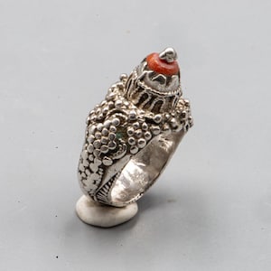Op de afbeelding: Een zilveren ring met een ingewikkelde, getextureerde band en een verhoogd, koepelvormig middelpunt bekroond met een kleine, ronde, oranjerode steen. Het metalen oppervlak vertoont gedetailleerde granulatie en filigraanachtige patronen over de band.