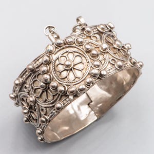 May include: Silver cuff bracelet with detailed floral and dot patterns. The hinged bracelet features intricate filigree work and small, raised spherical accents. The design includes multiple flower motifs and a clasp closure.