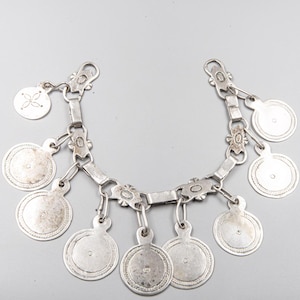 Puede incluir: Un collar o cinturón plateado con una serie de colgantes circulares y eslabones decorativos. Los colgantes tienen un punto central y un borde con dibujos. La pieza es probablemente vintage o antigua.
