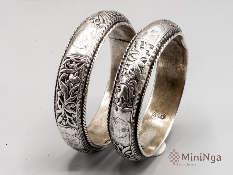 May include: Two silver bracelets with intricate floral and geometric designs. The bracelets are made of silver and have a polished finish.