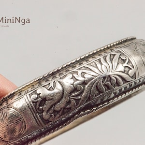 May include: Silver bangle bracelet with intricate floral and geometric designs. The bracelet is engraved with a repeating pattern of flowers and leaves.