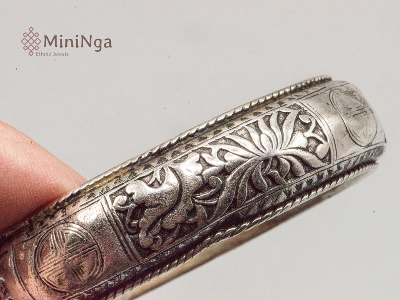 May include: Silver bangle bracelet with intricate floral and geometric designs. The bracelet is engraved with a repeating pattern of flowers and leaves.