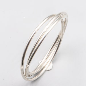 Può includere: Un braccialetto a cerchio color argento con tre fasce intrecciate. Le fasce sono lisce e rotonde, creando un design minimalista ed elegante. Il braccialetto è mostrato su una superficie bianca.