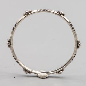Puede incluir: Un brazalete circular de plata con un acabado metálico pulido. La banda presenta cuatro grupos de pequeñas esferas de plata espaciadas uniformemente y secciones alternas de líneas negras diagonales grabadas en la superficie.