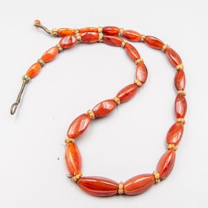 Peut inclure: Un collier composé de perles ovales rouge-orangé, séparées par de petits espaceurs beiges. Le collier est doté d'un fermoir en métal fin et foncé et d'une petite perle orange à l'extrémité. Les perles sont enfilées.