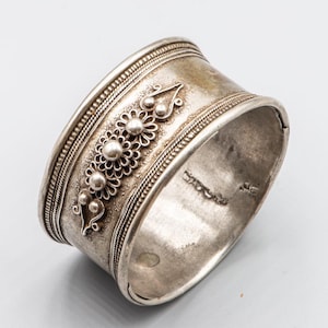 Peut inclure: Un bracelet manchette argenté orné d'un motif floral. Le bracelet large présente des détails complexes et des éléments floraux en relief. Les bords sont décorés de fines lignes. Le métal a un aspect légèrement vieilli.