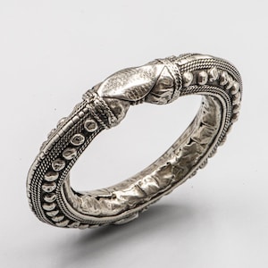 Può includere: Un braccialetto rigido argentato con dettagli intricati. Il braccialetto presenta una superficie strutturata con una serie di piccoli disegni circolari in rilievo e accenti simili a corde. Il design include un elemento centrale stilizzato. Diametro: circa 7 cm.