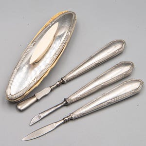 Puede incluir: Un juego de manicura de plata con tres herramientas y un estuche con un inserto blanco de forma ovalada. Las herramientas tienen mangos de plata adornados.