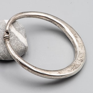 Puede incluir: Un brazalete ovalado plateado con una superficie texturizada. El brazalete tiene un diseño simple y elegante y parece estar hecho de metal. Los extremos del brazalete se conectan con un pequeño cierre decorativo. El brazalete está sobre una piedra gris.