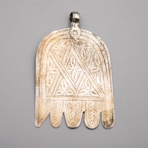 May include: A silver-colored pendant with intricate etched patterns. The pendant has a rounded top with a loop for hanging and a stylized hand shape with detailed designs. The surface is covered in geometric and floral motifs.