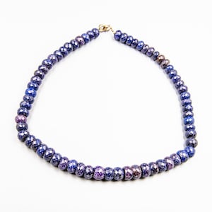 Peut inclure: Un collier composé de perles facettées bleu foncé et violettes. Les perles sont enfilées ensemble, formant un cercle complet. Le collier est sécurisé par un fermoir doré. Les perles ont un aspect brillant et poli.