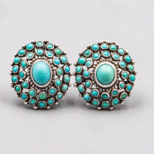 Può includere: Un paio di orecchini circolari in argento con un grande cabochon ovale di turchese al centro, circondato da molteplici pietre di turchese rotonde più piccole disposte a grappolo. Il metallo ha una finitura lucida con dettagli in perline d'argento.