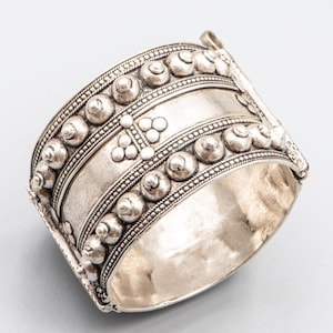 Puede incluir: Un brazalete ancho de tono plateado con detalles intrincados. El brazalete presenta filas de pequeños diseños circulares en relieve y patrones decorativos. El metal tiene una apariencia ligeramente envejecida, lo que sugiere un estilo vintage o antiguo. El brazalete está diseñado para usarse alrededor de la muñeca.