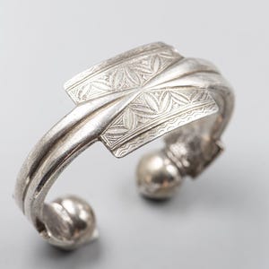May include: Silver cuff bracelet with intricate floral and geometric patterns. The bracelet features a rectangular centerpiece with detailed engravings and rounded ends with small, spherical accents. The metal has a polished, reflective finish.