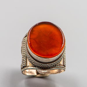 Puede incluir: Un anillo de plata con una piedra ovalada de color rojo anaranjado. El anillo tiene una banda detallada y texturizada con un patrón decorativo. La piedra está engastada en un bisel de plata, una joya.