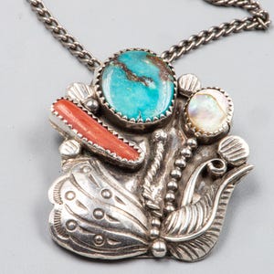 Puede incluir: Un collar con colgante de plata con un diseño de mariposa que incorpora turquesa, coral y nácar. El colgante tiene una superficie texturizada y detallada y cuelga de una cadena de plata. Este collar es una joya.