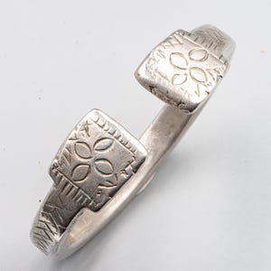May include: A silver-toned cuff bracelet with square terminals. Each terminal features an etched floral design within a square frame. The bracelet has an open design and is likely made of metal.