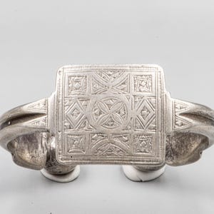 May include: A silver-toned ring with an engraved square centerpiece featuring geometric patterns. The ring's band curves outward, and the design includes diamond and cross-like shapes. The ring is likely made of metal.