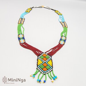 Op de afbeelding: Meerstrengige kralenketting met een geometrische hanger. De ketting is gemaakt van rode, groene, blauwe, gele en oranje kralen. De hanger heeft een ruitvorm met een franje van groene kralen.