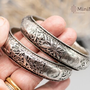 May include: Two silver bangles with intricate floral designs. The bangles are stacked on top of each other and are being held by a hand.