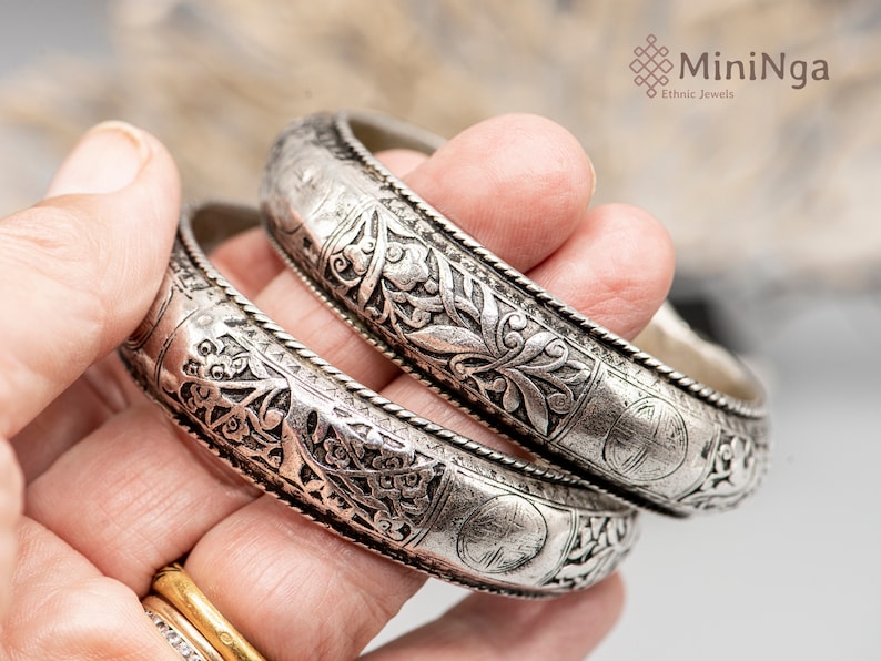 May include: Two silver bangles with intricate floral designs. The bangles are stacked on top of each other and are being held by a hand.