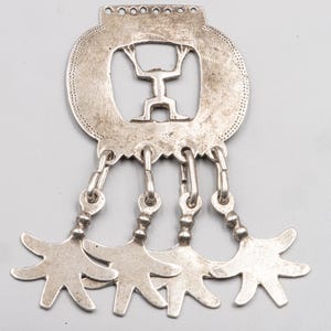 May include: A silver pendant with a central cutout figure holding up a weight. The pendant has a circular design with a dotted border and four dangling star-shaped charms. The pendant is made of a shiny metal.