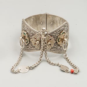 May include: Silver-toned cuff bracelet with intricate floral and scrollwork designs. The bracelet features a hinged design with decorative accents of red and green enamel. Two chains with small charms and a red bead dangle from the bracelet.