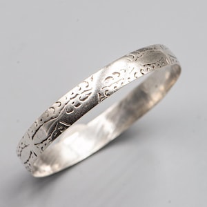 Może przedstawiać: Srebrzysta metalowa bransoletka typu bangle z ciągłym tłoczonym wzorem powtarzających się zakrzywionych kształtów i linii na zewnętrznej powierzchni. Obręcz ma gładkie, polerowane wnętrze oraz lekko zaokrąglony profil z metalicznym wykończeniem.