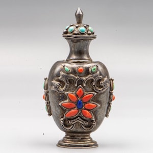 Op de afbeelding: Een snuiffles van zilverkleurig metaal met een afneembaar deksel met turkoois inleg. Het lichaam is versierd met een centraal bloemmotief van oranje en blauwe stenen, omringd door ingewikkeld metalen krulwerk en extra turkooiskleurige accenten.