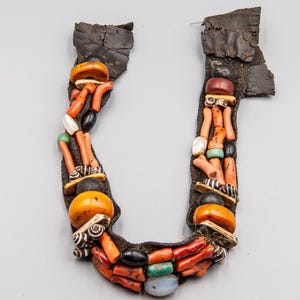 May include: A handcrafted necklace featuring a dark brown backing adorned with a variety of beads. The necklace incorporates orange, red, black, white, and green beads, creating a unique and colorful design. The necklace has a U-shape.