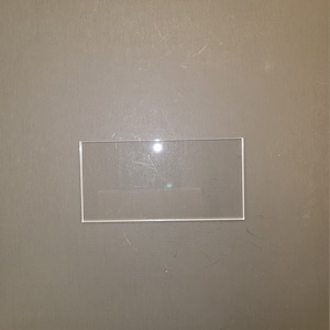May include: A clear, rectangular glass object with a thin, white border. The glass is positioned on a dark gray surface. A small, green dot is visible on the glass.