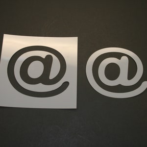 May include: Two white stencils of the "@" symbol, one on a white sheet of paper and the other on a black surface.